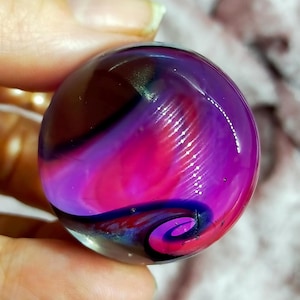 May include: A glass marble featuring a blend of purple, pink, and black swirling patterns. The marble has a glossy surface and a spiral design at the base. The colours merge, producing a distinctive and artistic look.
