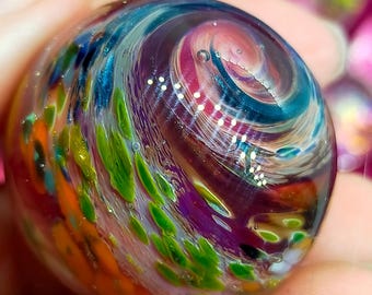 Pode incluir: Uma bola de gude de vidro colorida com um design em espiral. A bola de gude apresenta uma mistura de cores, incluindo azul, verde, laranja e roxo. O design assemelha-se a uma galáxia ou nebulosa.