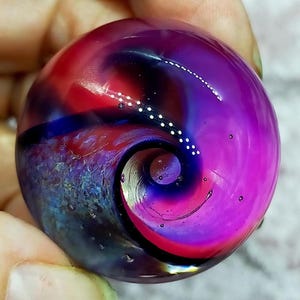 May include: A circular glass marble featuring a swirling pattern of deep purple, vibrant magenta, and dark blue hues. The surface is smooth and polished, containing small internal air bubbles and a distinct spiral design with white dots.