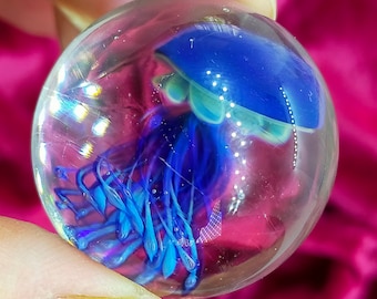May include: A clear glass marble with a blue jellyfish design. The jellyfish has a blue bell and flowing tentacles. The marble is held between fingers against a pink background. The jellyfish design is detailed and appears to be suspended within the glass.