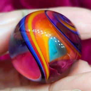 May include: A spherical glass marble featuring a vibrant swirl pattern of translucent orange, deep purple, bright yellow, and cyan blue colors. The surface is smooth and polished, reflecting light with distinct white highlights across the curved glass.