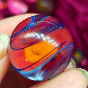 May include: A spherical glass marble featuring a vibrant orange core surrounded by layers of translucent red glass. The surface is decorated with swirling dark blue ribbons and contains small internal air bubbles throughout the clear glass structure.