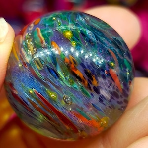 May include: A colorful glass marble with swirling patterns of blue, green, orange, and red. The marble has a glossy finish and small, golden-yellow bubbles throughout. The overall effect is a vibrant and artistic piece.