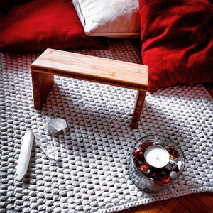 Könnte beinhalten: Eine Holz-Meditationsbank mit einem weißen, strukturierten Teppich darunter. Es gibt zwei klare Kristalle und ein Glasgefäß mit roten und weißen Dekorationen und einer kleinen weißen Kerze.