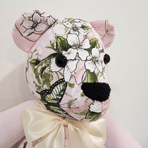 May include: A pink teddy bear with a floral print and a butterfly design. The bear has black eyes and a black nose. It is wearing a cream-colored satin bow.