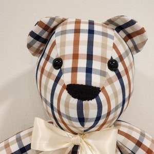 May include: A plaid teddy bear with a cream-colored satin bow. The bear is made of a brown, white, and blue plaid fabric. The bear has black button eyes and a black felt nose.