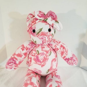 May include: A handmade plush bear with a floral rose pattern in shades of pink and white. The bear has black button eyes and a matching floral bow on its head. The arms and legs are attached, and the bear is sitting upright.