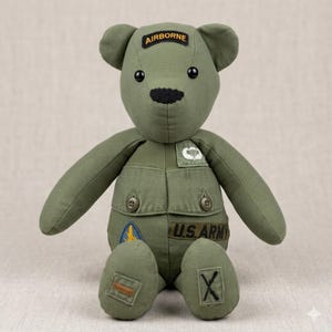May include: A handmade olive green teddy bear wearing a military-style uniform. The bear has black button eyes, a black stitched nose, and patches that read "AIRBORNE" and "U.S. ARMY". The bear is sitting with its arms and legs extended.