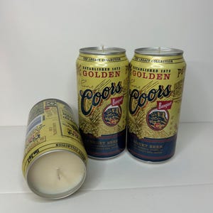 May include: Three Coors Banquet beer can candles. The candles are in the shape of the beer cans, with the original branding and text. The cans are yellow with blue and red accents. One candle is lying on its side, revealing the candle wax.