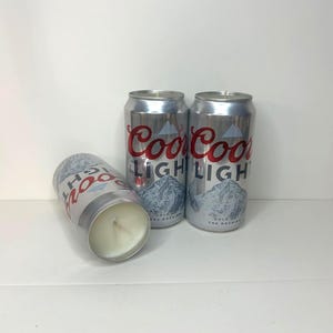 May include: Three silver-colored candles shaped like beer cans. The cans have the words "Coors Light" in red and a mountain graphic. One candle is lying on its side, revealing the white wax and wick.