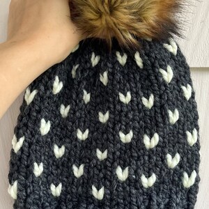 May include: A black knit beanie with a white heart pattern and a brown faux fur pom pom. The beanie has a brown leather tag with the word "freya" on it.