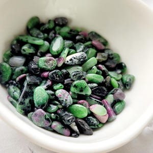 May include: A white ceramic bowl filled with polished gemstones. The stones are a mix of green, black, and pink hues. The gemstones are oval and irregular in shape. The bowl is on a white surface with clear crystal accents.