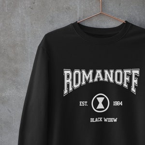 May include: Black sweatshirt with white text reading "ROMANOFF", a circular graphic, and the words "EST. 1984 BLACK WIDOW". The garment hangs on a copper-coloured hanger against a grey wall.