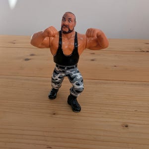 May include: A plastic action figure of a wrestler with a shaved head and beard, wearing a black tank top, grey and white camouflage trousers, and black boots. The figure is posed with both arms raised in a flexing position with clenched fists.