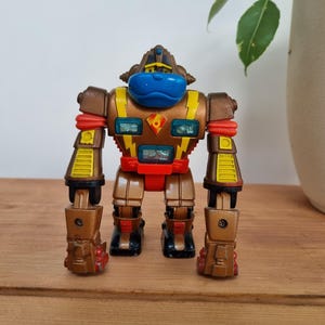 Puede incluir: Un robot de juguete vintage con cuerpo marrón y dorado, cara azul y detalles rojos. El robot tiene un diseño detallado con articulaciones visibles y una cabeza que se asemeja a un gorila. Está sobre una superficie de madera.