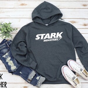 May include: Dark heather grey hoodie with a white graphic of the Stark Industries logo. The logo features the word "Stark" in a bold white font with a white arrow pointing to the right. The words "Industries" are printed in a smaller white font below the word "Stark".