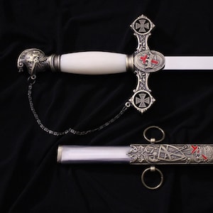 May include: A decorative sword with a white handle and blade, featuring ornate silver-colored metalwork. The hilt includes a cross design with red accents. A matching scabbard is also present, with similar detailing and rings.