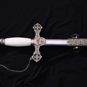 May include: An ornate sword with a white handle and silver accents. The blade is silver, and the hilt features a cross design with red and silver details. A chain is attached to the pommel.