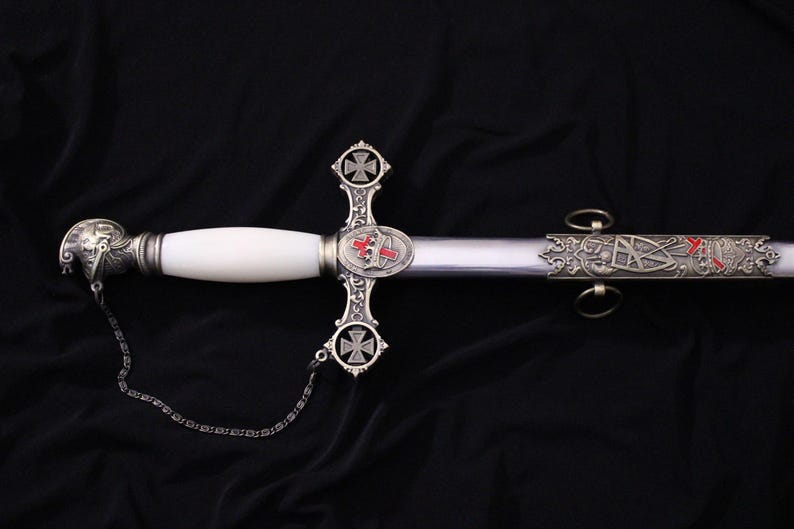 May include: An ornate sword with a white handle and silver accents. The blade is silver, and the hilt features a cross design with red and silver details. A chain is attached to the pommel.