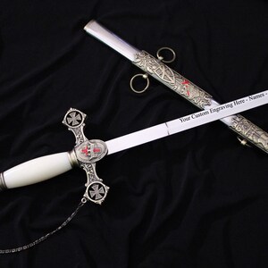 May include: A ceremonial sword with a white handle and a silver blade. The hilt features a cross design with red accents. The scabbard is silver with ornate detailing. Text on the blade reads "Your Custom Engraving Here - Names - Dates - Details."