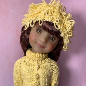 May include: A doll in a handmade yellow knitted dress and matching hat. The dress has a turtleneck and flower-shaped buttons. The doll is wearing gold sandals. The background is a pale pink.