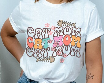 Könnte beinhalten: Weißes T-Shirt mit dem Text "Cat Mom Cat Mom Cat Mom" in einer Retro-Schriftart mit bunten Buchstaben, Pfotenabdrücken und Fischgrätenmustern.