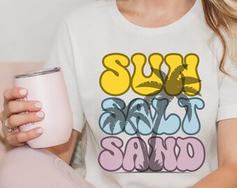 Könnte beinhalten: Weißes T-Shirt mit einem Grafikdesign, das "SUN SALT SAND" in bunten, Retro-Lettern mit Palmen zeigt.