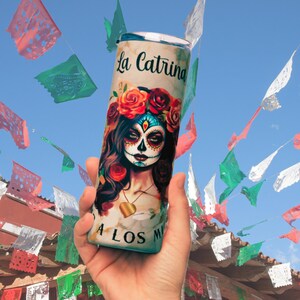 May include: A white and gold tumbler with a colorful illustration of a sugar skull with the text "La Catrina A Los Muertos".