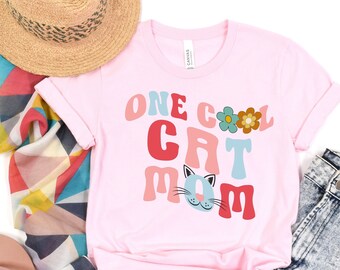 Könnte beinhalten: Pinkes T-Shirt mit dem Text "One Cool Cat Mom" in bunter Retro-Schrift. Der Text ist von Blumen und einem Katzenkopf umgeben.