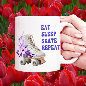 May include: White ceramic mug with a purple floral design and a white roller skate with purple accents. The text "Eat Sleep Skate Repeat" is printed in purple on the mug.
