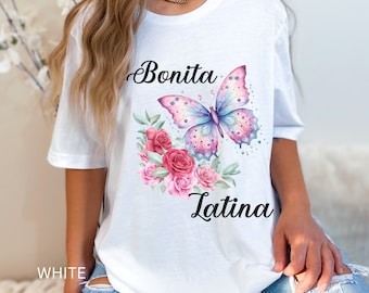 Könnte beinhalten: Weißes T-Shirt mit einem rosa und violetten Schmetterling und Blumen. Der Text "Bonita Latina" ist auf das T-Shirt gedruckt.