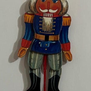 Op de afbeelding: Een houten kerstversiering in de vorm van een notenkraker met een rood, wit en blauw uniform, een zwarte hoed met een wit hakbijlontwerp en een witte baard. De notenkraker is bevestigd aan een rood touwtje met een rode houten bal aan het uiteinde.