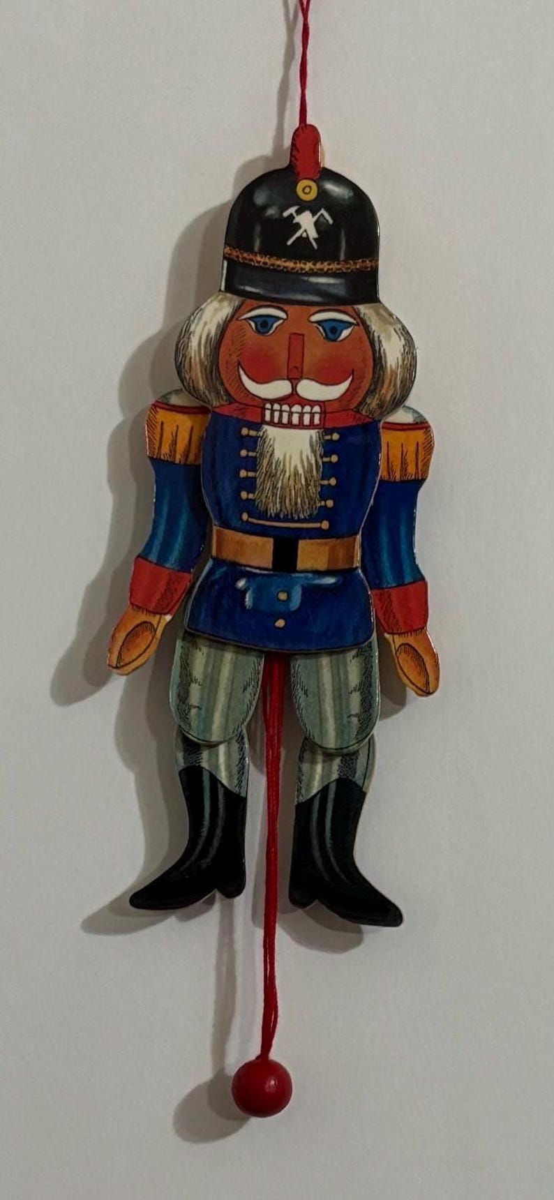 Op de afbeelding: Een houten kerstversiering in de vorm van een notenkraker met een rood, wit en blauw uniform, een zwarte hoed met een wit hakbijlontwerp en een witte baard. De notenkraker is bevestigd aan een rood touwtje met een rode houten bal aan het uiteinde.