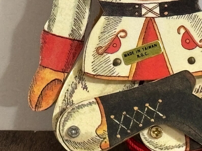 May include: A vintage wooden toy soldier with red, white, and black painted details. The soldier is wearing a black belt with a gold buckle and has a gold label that reads "Made in Taiwan R.O.C."