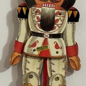May include: A red, white, and black wooden nutcracker doll with a painted face and a black mustache. The doll is wearing a white uniform with red and gold accents. The doll is holding a string in its hand. The doll is labeled "Made in Taiwan R.O.C."