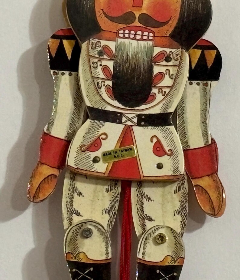 May include: A red, white, and black wooden nutcracker doll with a painted face and a black mustache. The doll is wearing a white uniform with red and gold accents. The doll is holding a string in its hand. The doll is labeled "Made in Taiwan R.O.C."