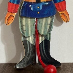 Op de afbeelding: Een houten speelgoed soldaat met een blauw en rood uniform, gestreepte broek en zwarte laarzen. De soldaat is bevestigd aan een rood touw met een rode houten bal aan het einde.