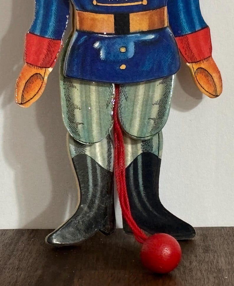 Op de afbeelding: Een houten speelgoed soldaat met een blauw en rood uniform, gestreepte broek en zwarte laarzen. De soldaat is bevestigd aan een rood touw met een rode houten bal aan het einde.