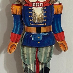 Op de afbeelding: Een houten kerstversiering in de vorm van een notenkraker met een rood, wit en blauw uniform. De notenkraker heeft een zwarte hoed met een pikhouwelsymbool, een witte baard en een rode bal aan een touwtje.