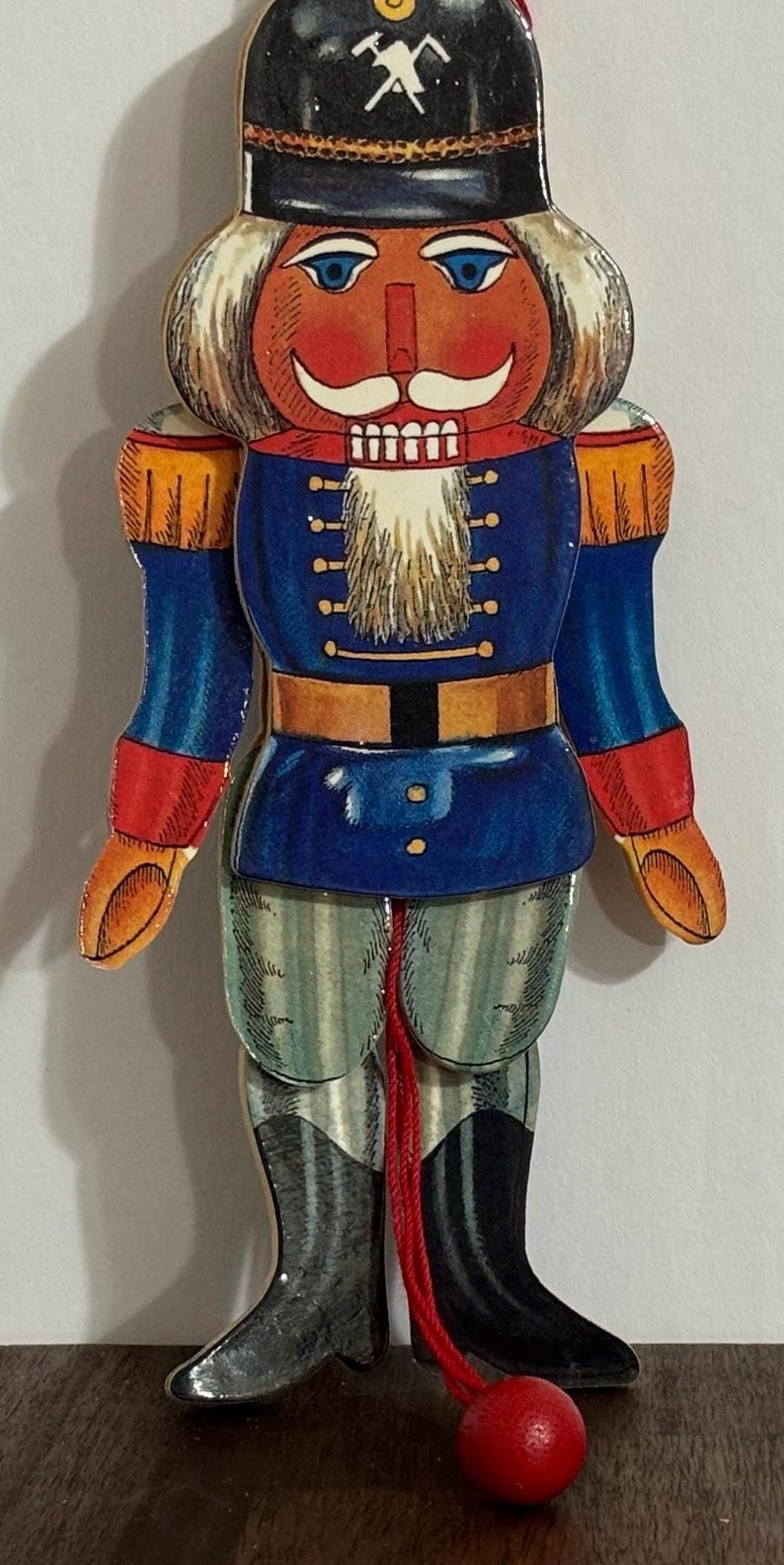 Op de afbeelding: Een houten kerstversiering in de vorm van een notenkraker met een rood, wit en blauw uniform. De notenkraker heeft een zwarte hoed met een pikhouwelsymbool, een witte baard en een rode bal aan een touwtje.
