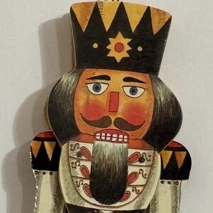 May include: A vintage wooden nutcracker ornament with a red, white, and black painted design. The nutcracker has a black crown, a white beard, and a red nose. The ornament is made in Taiwan.