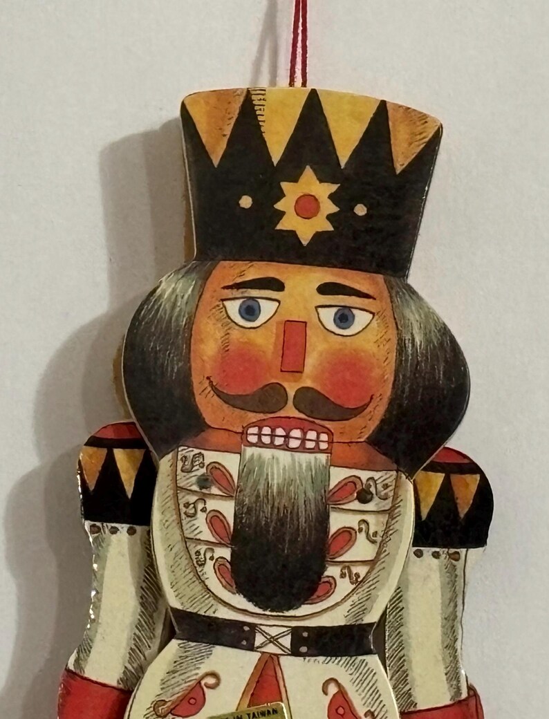 May include: A vintage wooden nutcracker ornament with a red, white, and black painted design. The nutcracker has a black crown, a white beard, and a red nose. The ornament is made in Taiwan.