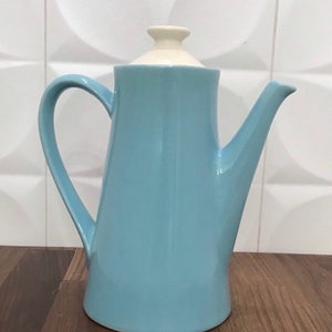 以下が含まれることがあります： 白い蓋が付いた薄い青色のセラミック製のコーヒーポット。ポットは丸みを帯びたボディと側面に注ぎ口があります。