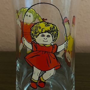 May include: A clear glass drinking glass with a red, yellow, and white illustration of three Cabbage Patch Kids dolls jumping rope. The dolls are wearing red dresses and yellow shoes.