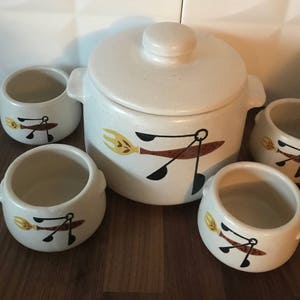 Puede incluir: Conjunto de cinco cuencos de cerámica blanca con diseños pintados en marrón y amarillo. Los cuencos presentan una imagen estilizada de un tenedor y una cuchara. El cuenco más grande tiene una tapa.