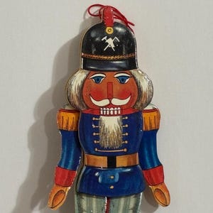May include: A wooden nutcracker ornament with a red, white, and blue uniform. The nutcracker has a white beard, a red face, and a black hat with a gold pickaxe symbol.