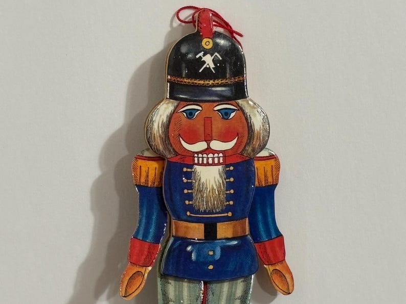 Op de afbeelding: Een houten notenkraker ornament met een rood, wit en blauw uniform. De notenkraker heeft een witte baard, een rood gezicht en een zwarte hoed met een gouden houweel symbool.