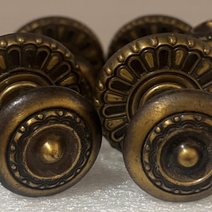 May include: A set of round brass cabinet knobs featuring a dark, aged finish. Each knob has a central raised dome surrounded by a decorative border with a scalloped, petal-like pattern and a beaded edge, providing a textured, metallic appearance.