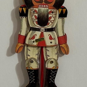 May include: A red, white, and black wooden nutcracker ornament with a red string and a red ball at the end. The nutcracker is wearing a white and red uniform with a black belt and a black crown. The nutcracker has a black beard and mustache. The ornament is labeled "Made in Taiwan K.C."