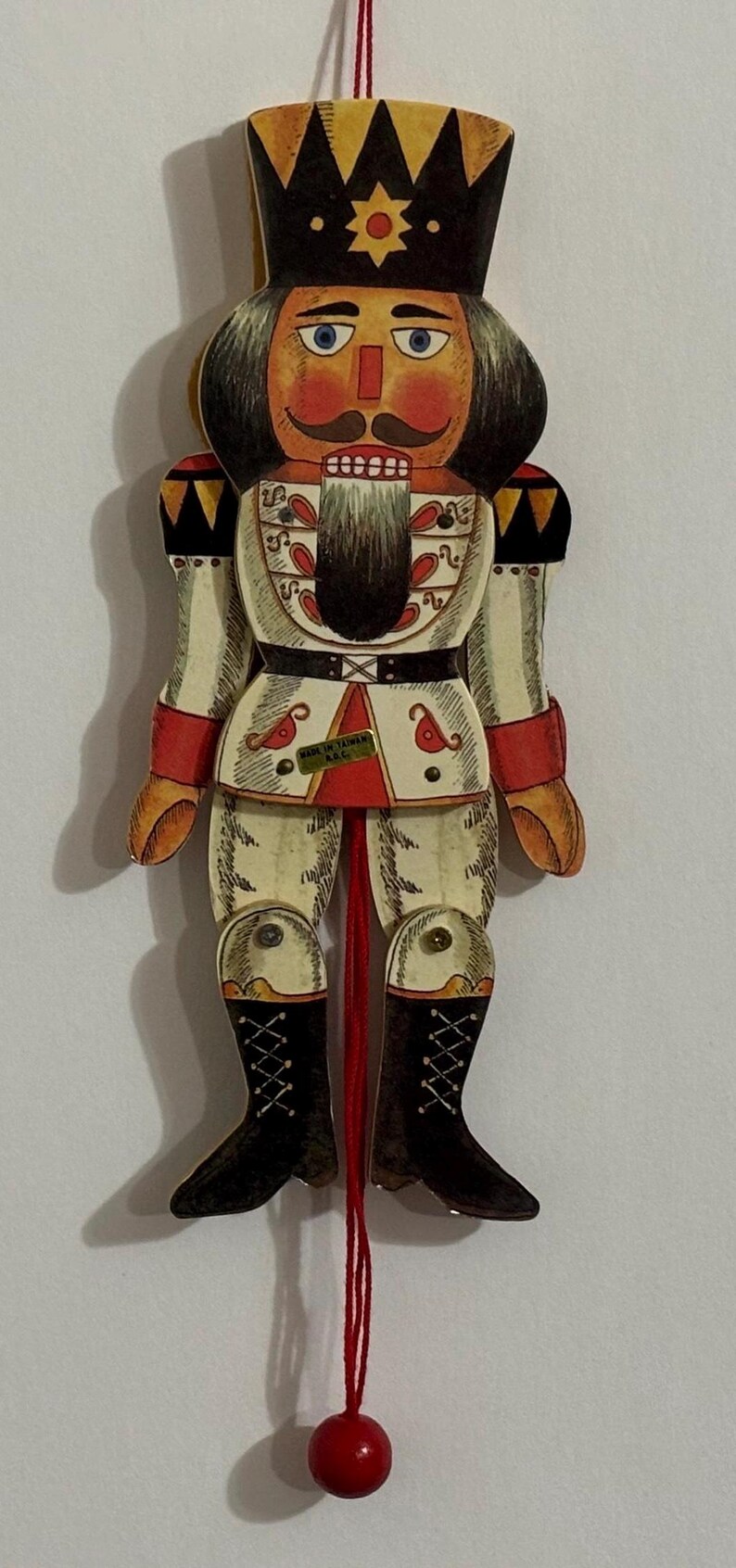 May include: A red, white, and black wooden nutcracker ornament with a red string and a red ball at the end. The nutcracker is wearing a white and red uniform with a black belt and a black crown. The nutcracker has a black beard and mustache. The ornament is labeled "Made in Taiwan K.C."